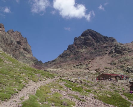 A hut with people standing outside it, with grassy and rocky ground and stoney hills in the background