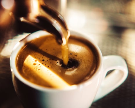 An espresso shot pours from the metal spout of an espresso machine into a small white cup of espresso