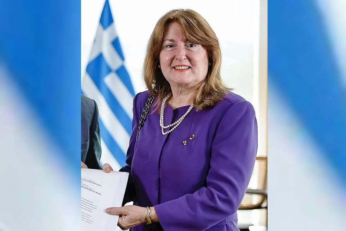 Mulher de terno lilás com uma bandeira da Grécia ao fundo. Trata-se da embaixadora da Grécia, Eleni Lianidou - Metrópoles